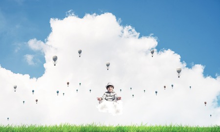 Young little boy keeping eyes closed and looking concentrated while meditating on cloud among flying aerostats with bright and beautiful landscape on background.の写真素材