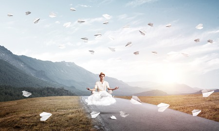 Young man keeping eyes closed and looking concentrated while meditating on cloud above the road with beautiful and breathtaking landscape on background.の写真素材