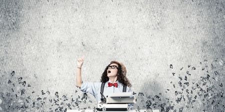 Young and beautiful woman writer in hat and eyeglasses using typing machine and pointing upside while sitting at the table among flying letters and against gray concrete wall on background.の写真素材