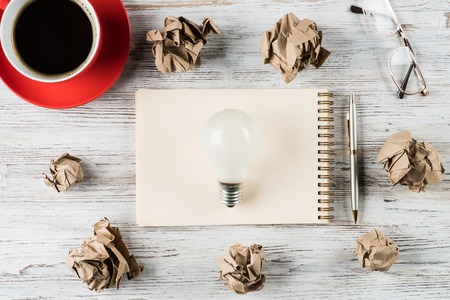 Top view of modern creative workplace with lightbulb on notepad, pen, crumpled paper balls and coffee on wooden table. Concept of finding of outstanding idea.の写真素材