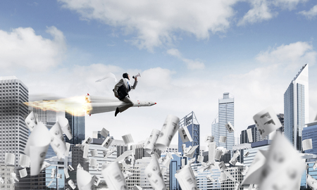 Conceptual image of young and happy businessman in suit flying on rocket among flying papers with modern cityscape with skyscrapers and blue sky on background.の写真素材