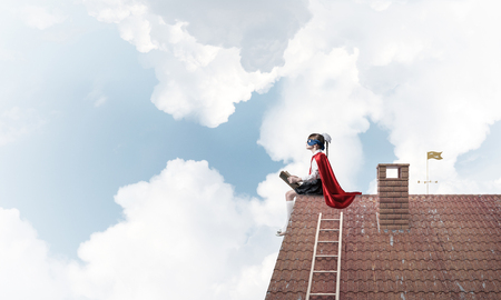 Little child in mask and cape sitting on house brick roof and reading. Mixed mediaの写真素材