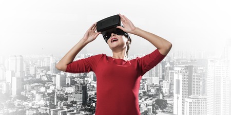 Horizontal shot of young and beautiful woman in red dress using virtual reality headset while standing outdoors with modern cityscape view on backgroundの写真素材