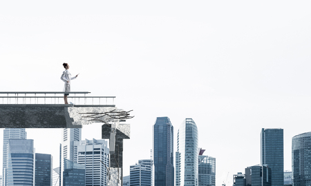 Conceptual image of woman doctor in white sterile suit standing on edge of broken bridge with cityscape view on background. Medical industry conceptの写真素材