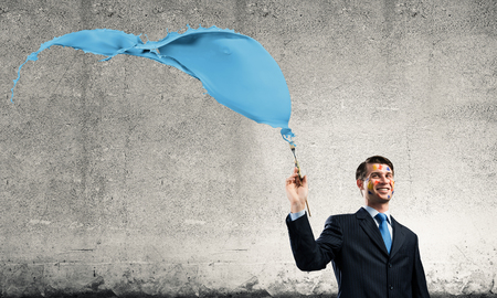 Horizontal shot of cheerful and young businessman in black suit standing with blue coloured splash against dark gray wall on background.の写真素材