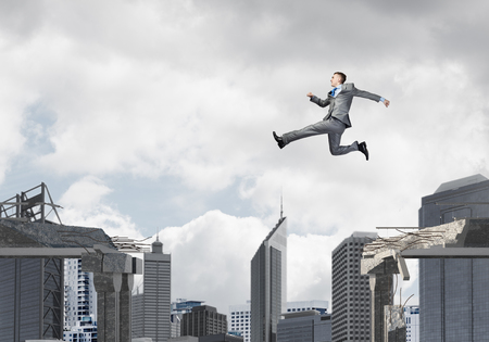 Businessman jumping over gap in concrete bridge as symbol of overcoming challenges. Dark sky and cityscape on background. 3D rendering.の写真素材