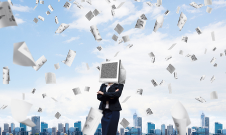 Cropped image of business woman in suit with monitor instead of head keeping arms crossed while standing outdoors among flying papers with cityscape view on background.の写真素材