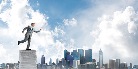 Businessman in suit running with phone in hand on pile of documents with cityscape on background. Mixed media.の写真素材