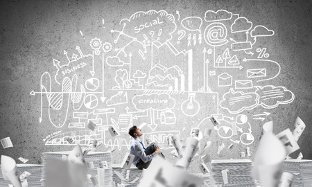 Businessman looking away while sitting on pile of documents among flying papers with business-analytical information on background. Mixed media.の写真素材