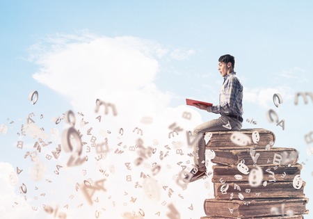 Young shocked man in casual sitting on pile of books with one in handsの写真素材