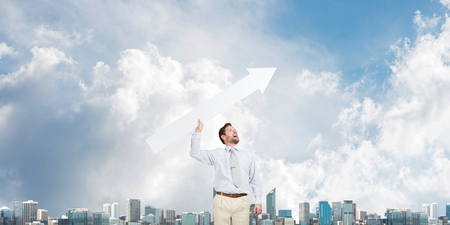 Horizontal shot of young businessman throwing big white arrow in the air while standing against modern cityscape view and cloudy sky on backgroundの写真素材