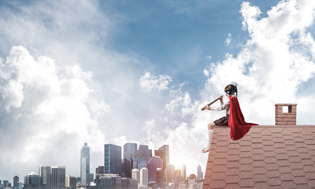 Little child in mask and cape sitting on house brick roof and looking in spyglass. Mixed mediaの写真素材