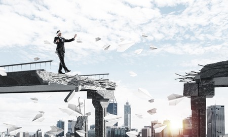 Businessman walking blindfolded among flying paper planes on concrete bridge with huge gap as symbol of hidden threats and risks. Cityscape and sunlight on background. 3D rendering.の写真素材