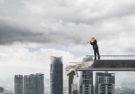 Young engineer in suit and helmet looking in spyglass while standing on broken bridge with cityscape on background. 3D rendering.の写真素材
