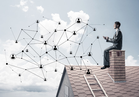 Young businessman sitting on house chimney with smartphone in handsの写真素材