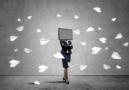 Business woman in suit with TV instead of head keeping arms crossed while standing among flying paper planes inside empty room with gray dark wall on background.の写真素材