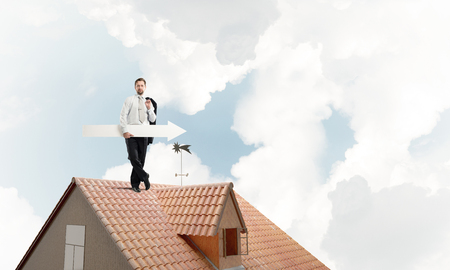 Young and ambitious businessman in suit holding big white arrow in hand and pointing away while standing on brick roof with cloudy skyscape view on background.の写真素材