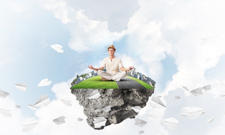 Man in white clothing keeping eyes closed and looking concentrated while meditating on island in the air among flying paper planes with cloudy skyscape on background. 3D rendering.の写真素材