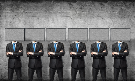 Businessmen in suits with TV instead of their heads keeping arms crossed while standing in a row in empty room with gray wall on background.の写真素材