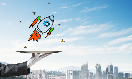 Cropped image of waitresss hand in white glove presenting sketched flying missile on metal tray with cityscape view on background.の写真素材