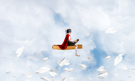 Little child in mask and cape floating on book in sky and readingの写真素材