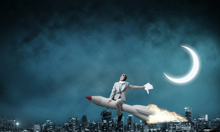 Conceptual image of young businessman in suit flying on rocket with night cityscape and blue sky on background.の写真素材