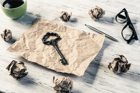 Top view of modern creative workplace with key on creased piece of sheet, crumpled paper balls and cup of coffee placed on wooden table. Concept of finding of outstanding idea.の写真素材