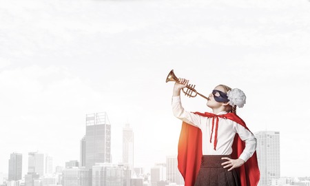 Little confident child in mask and cape plays trumpet against sunset backgroundの写真素材