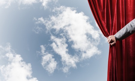 Human hand opens red velvet curtain on blue sky backgroundの写真素材