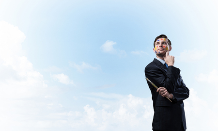 Portrait of young thoughtful man holding paintbrush in his hand and looking away while standing against blue cloudy skyscape on background.の写真素材