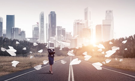 Business woman in suit with monitor instead of head keeping arms crossed while standing on the road among flying paper planes with beautiful landscape on background.の写真素材