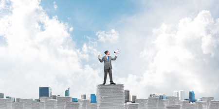 Businessman in suit standing on pile of documents with speaker in hand with skyscape and city view on background. Mixed media.の写真素材