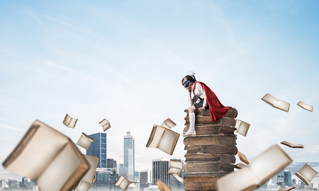 Little sad child in mask and cape sitting on pile of books and looking down. Mixed mediaの写真素材