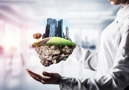 Closeup of business woman in shirt keeping green island with skycraper city in her hands with office view and sunlight on background. Mixed media.の写真素材