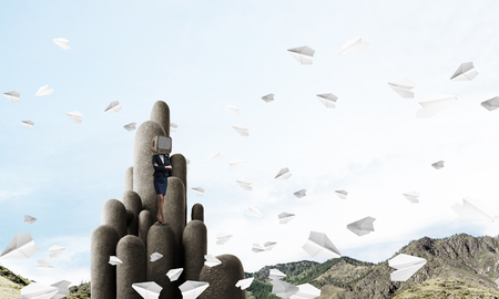 Business woman in suit with an old TV instead of head keeping arms crossed while standing on the top of stone columns among flying paper planes with beautiful landscape on background. 3D rendering.の写真素材