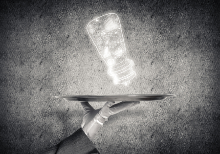 Closeup of waitresss hand in glove presenting sketched business plan in form of lamp on metal tray with gray wall on background.の写真素材