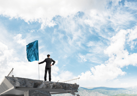 Rear view of confident businessman in suit holding flag in hand while standing on broken bridge with cloudly skyscape and nature view on background. 3D rendering.の写真素材