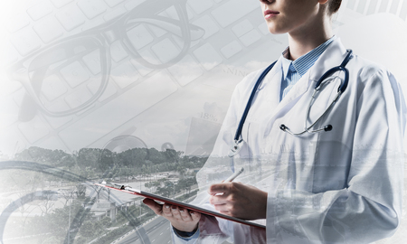 Portrait of young female doctor in white sterile suit holding notebook in hands while standing against modern city view and medical equipment on background. Double exposureの写真素材