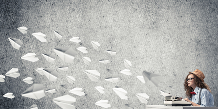 Young and beautiful woman writer in hat and eyeglasses using typing machine while sitting at the table among flying paper planes and against gray concrete wall on background.の写真素材