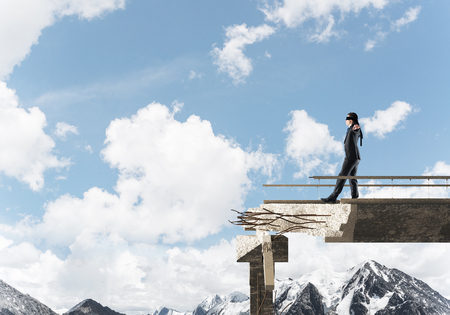 Businessman walking blindfolded on concrete bridge with huge gap as symbol of hidden threats and risks. Skyscape and nature view on background. 3D rendering.の写真素材