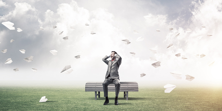 Frustrated businessman sitting on bench and closing ears with hands and paper planes fly aroundの写真素材