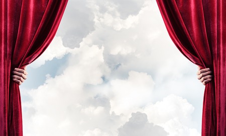 Human hand opens red velvet curtain on blue sky backgroundの写真素材