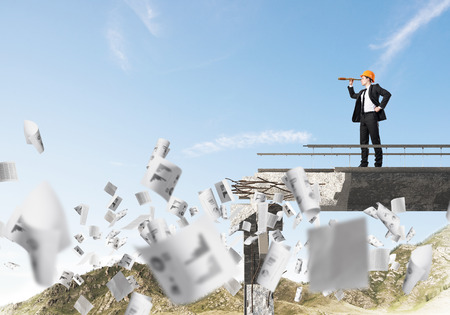 Young engineer in suit and helmet looking in spyglass while standing among flying papers on broken bridge with skyscape on background. 3D rendering.の写真素材