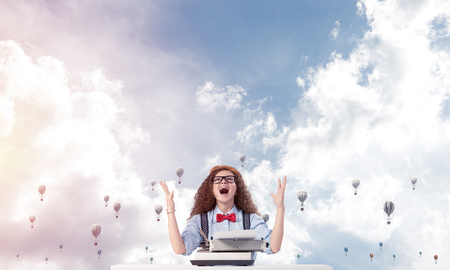 Young and beautiful woman writer in hat and eyeglasses using typing machine while sitting at the table with flying aerostats and cloudy skyscape on background.の写真素材