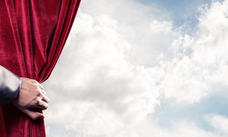 Human hand opens red velvet curtain on blue sky backgroundの写真素材