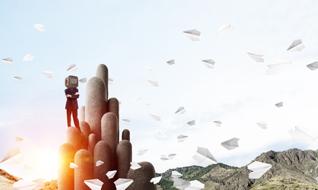 Businessman in suit with an old TV instead of head keeping arms crossed while standing on the top of stone columns among flying paper planes with beautiful landscape on background. 3D rendering.の写真素材