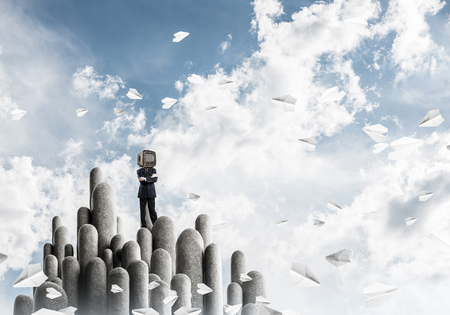 Businessman in suit with an old TV instead of head keeping arms crossed while standing on the top of stone columns among flying paper planes with beautiful landscape on background. 3D rendering.の写真素材