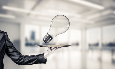 Cropped image of waitresss hand in white glove presenting lightbulb on metal tray as symbol of new idea with office view on background.の写真素材