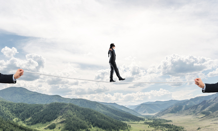 Businessman walking blindfolded on rope above high mountains as symbol of hidden threats and support. Skyscape and nature view on background.の写真素材