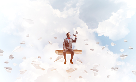 Young businessman sitting on book and screaming emotionally in megaphoneの写真素材